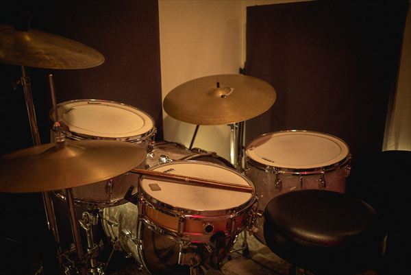 A drum kit with cymbals, a snare and a pair of sticks resting across it, lit warm against dark studio walls.