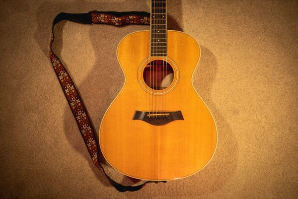 An acoustic guitar lying face up on the carpet with a patterned woven strap trailing off to the side.