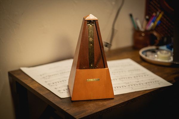 A wind-up wooden metronome standing on an open page of sheet music, with a jar of pencils behind it.
