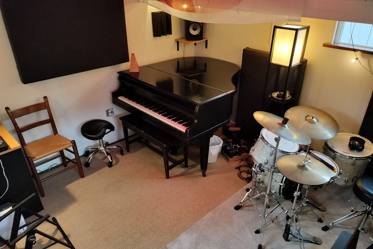 The studio's main room: a black grand piano against the wall, a drum kit in the corner, sound panels, studio monitors and a warm floor lamp.
